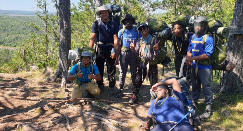 A group of students wearing backpacks pose for a photo in a wooded area. They appear to be at high elevation, as there are trees in the background below them.